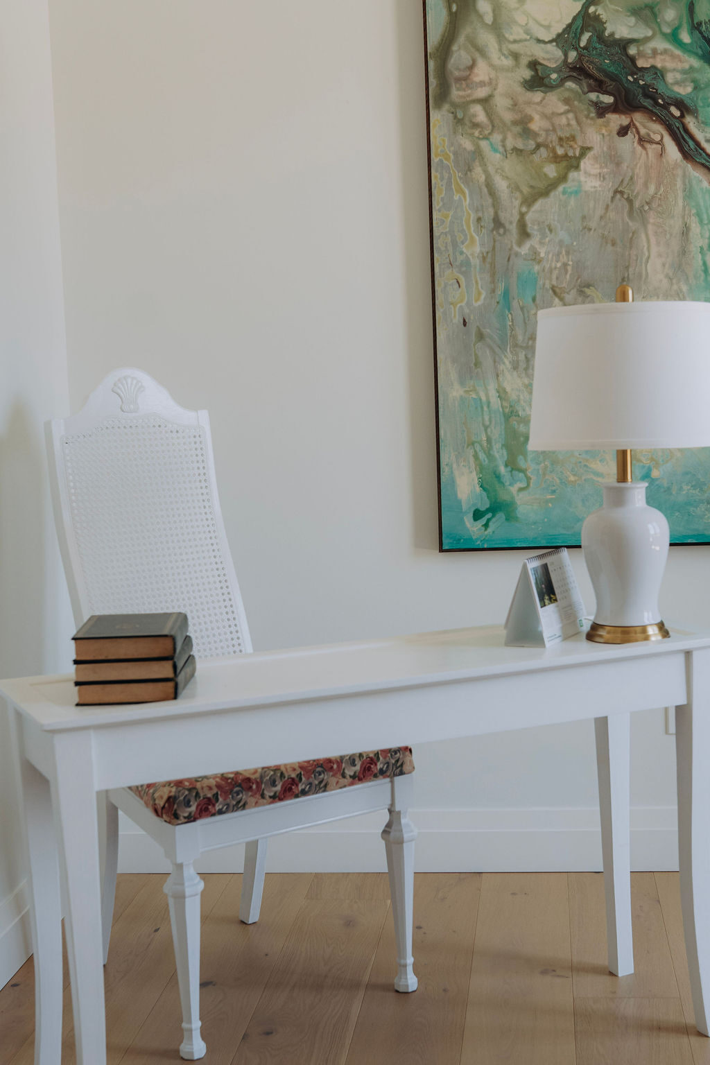 Entry console table in white with a cane-back chair, a stack of books and a lamp beneath framed artwork