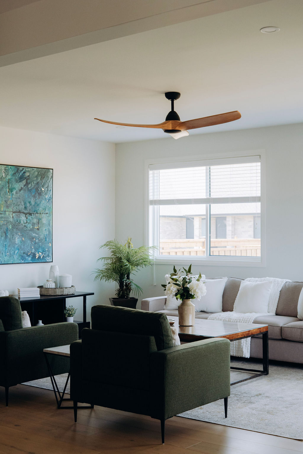 The great room of the model home, with a wood-blade ceiling fan, a cream sectional under the windows and an olive armchair beside a teal abstract canvas