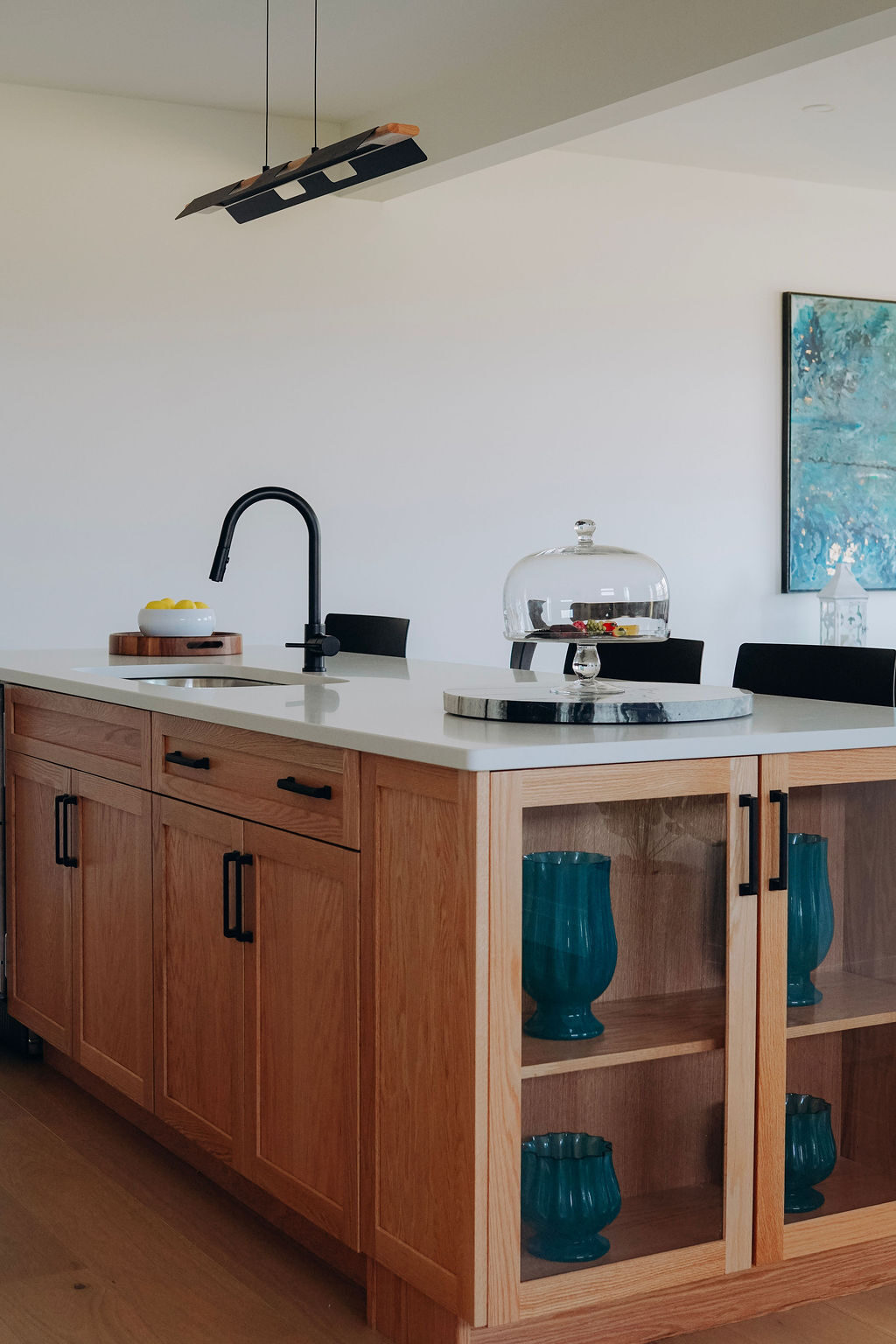 The oak island in quartz, with a black gooseneck faucet and open shelving holding teal glassware