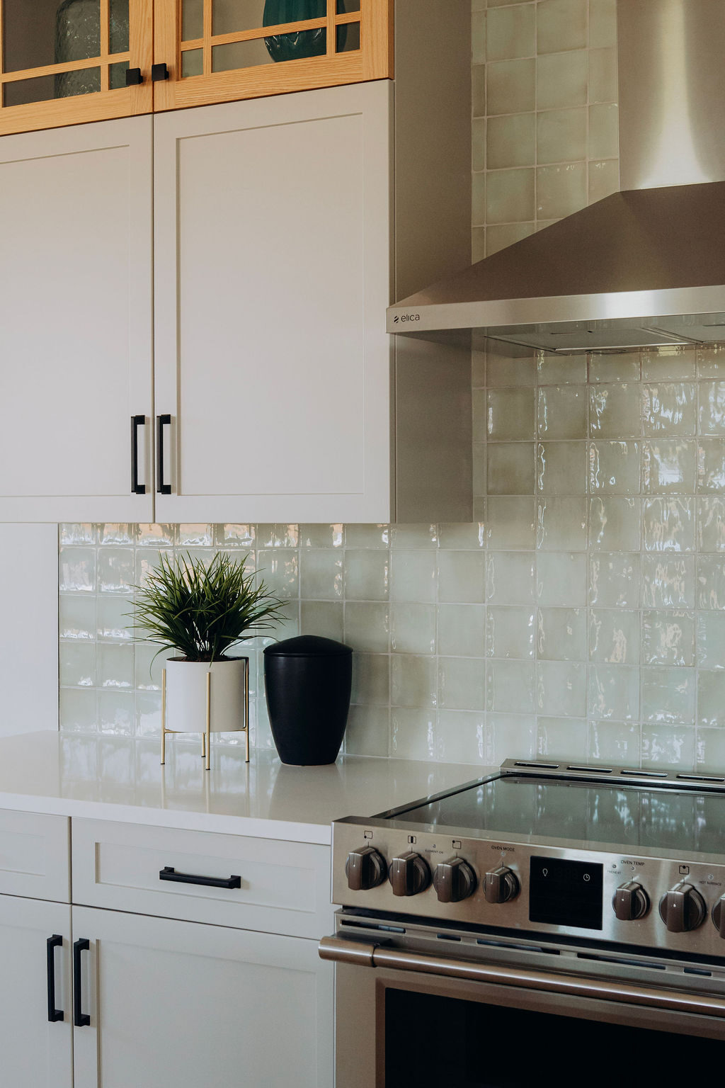 The range wall, with white shaker cabinets, glass-front oak uppers, a stainless hood and a pale green tiled backsplash