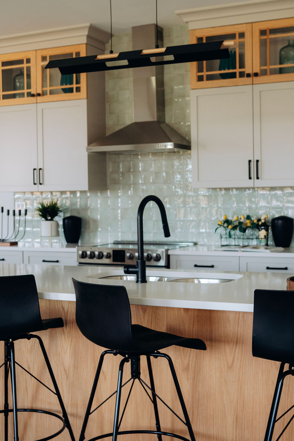 The model home kitchen, with an oak island and black stools under a linear pendant, glass-front oak upper cabinets and a stainless hood