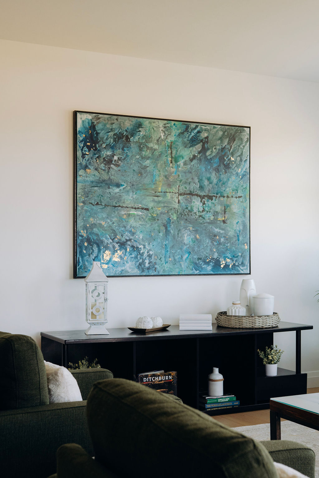 A large teal abstract canvas above a black console table, with olive armchairs in the foreground
