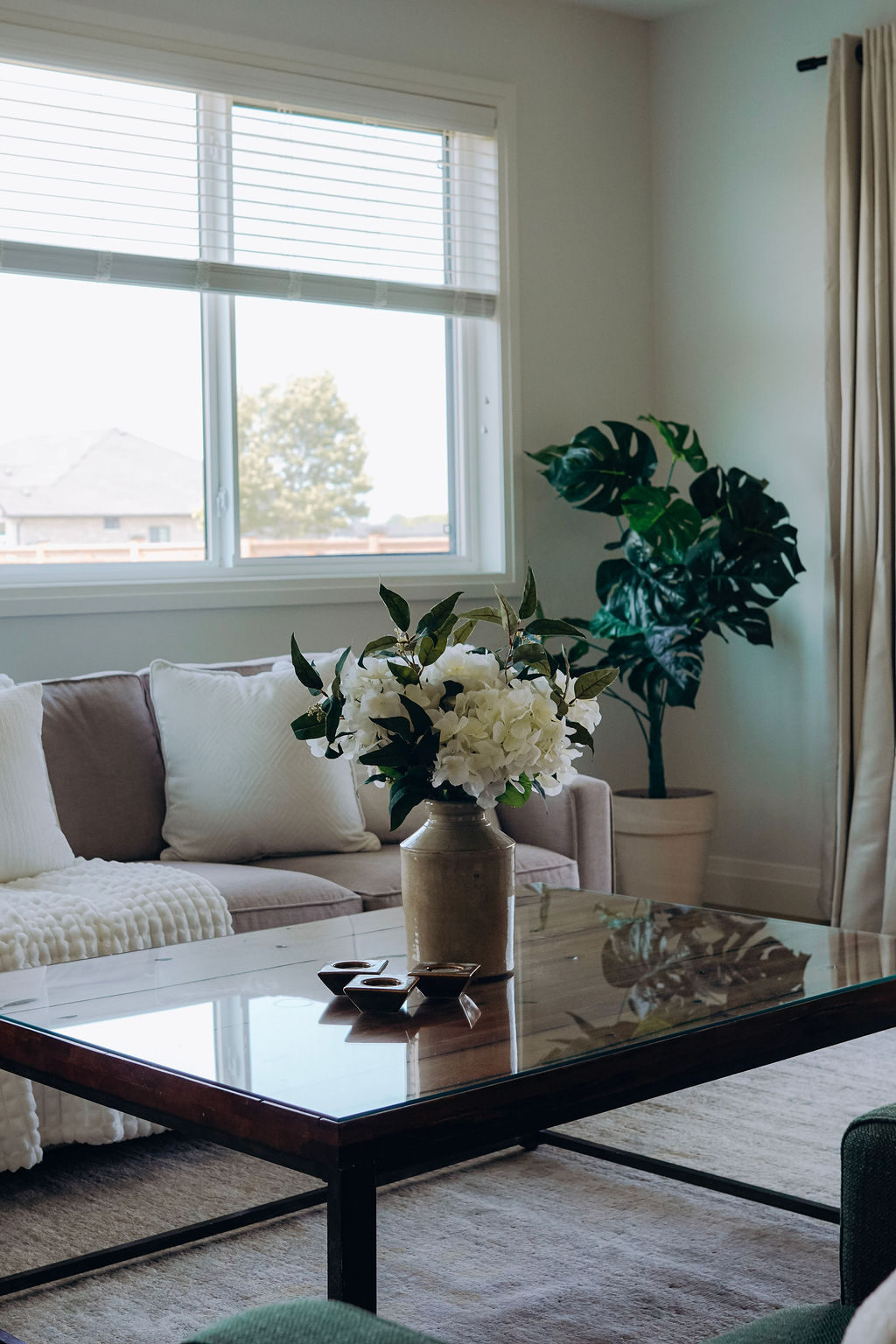 Living room seating beside a window in the model home, with hydrangeas on the glass-topped coffee table and a monstera in the corner